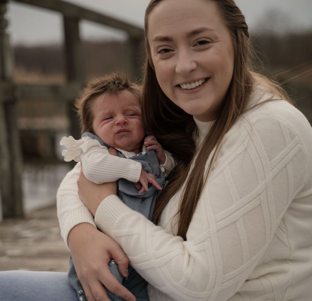 Smiling young mother in white sweater holding her newborn baby outdoors near a wooden bridge.