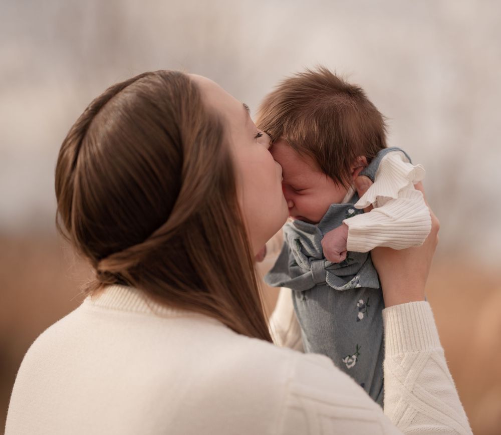 A young mother tenderly kisses her newborn baby while holding them up close outdoors.