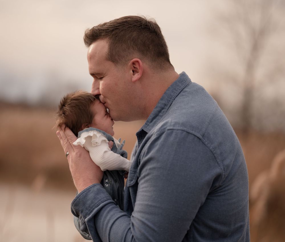 A father tenderly kisses his newborn baby outdoors near a lake in a heartwarming family portrait.