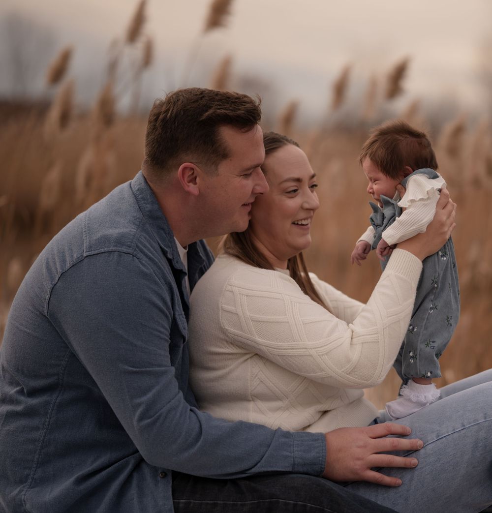 A happy couple smiles at their newborn baby outdoors in a golden field.