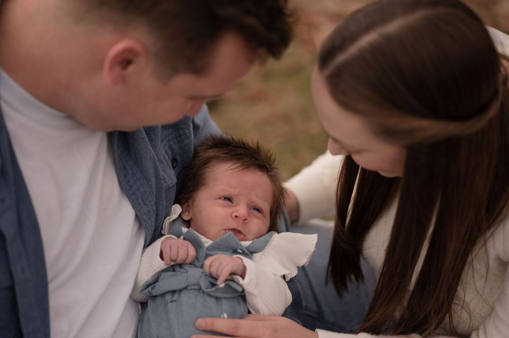 Parents lovingly gazing at their newborn baby dressed in a light blue outfit during an outdoor family photo session.