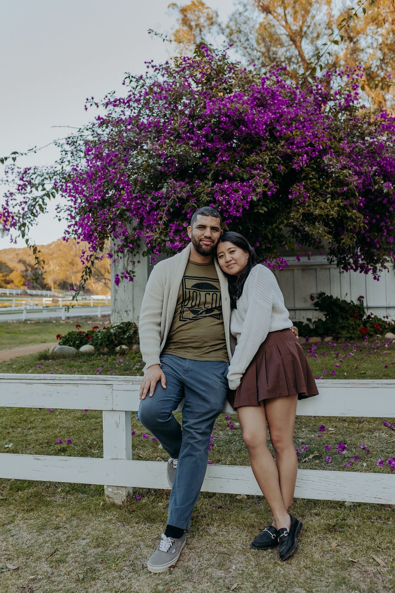 A couple leans against a white fence with vibrant purple bougainvillea flowers blooming behind them at Will Rogers State Park.