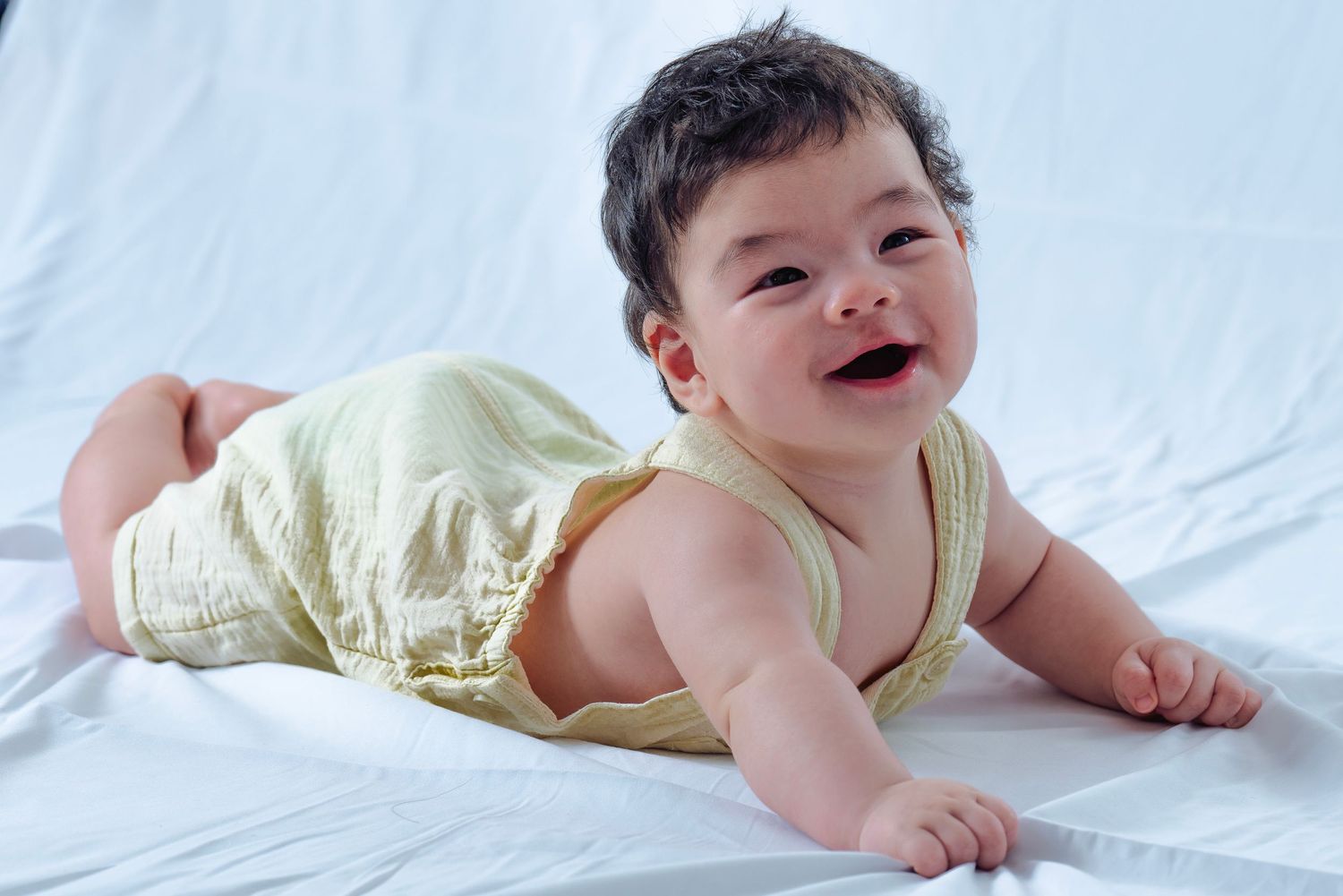 Baby in yellow outfit lying on white bedding with bright natural lighting.