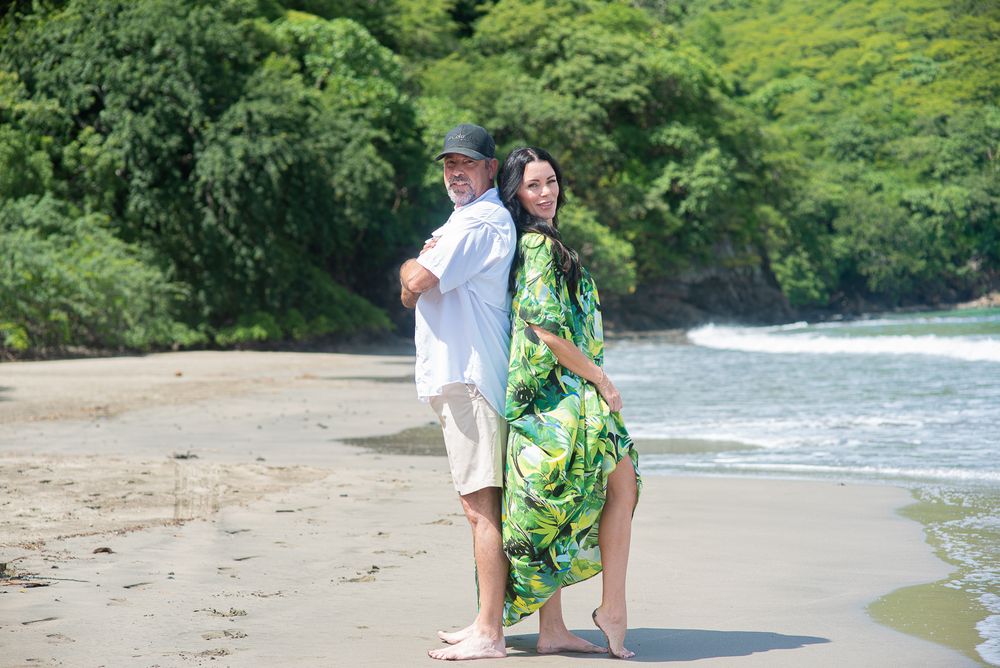 A person in a green floral dress walks along a scenic tropical beach with waves and lush vegetation in the background.