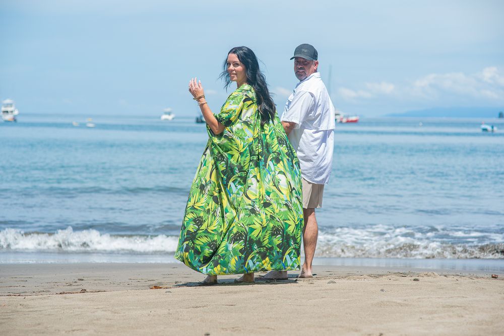 Couple walking along a scenic beach with blue ocean waters and boats in the background.