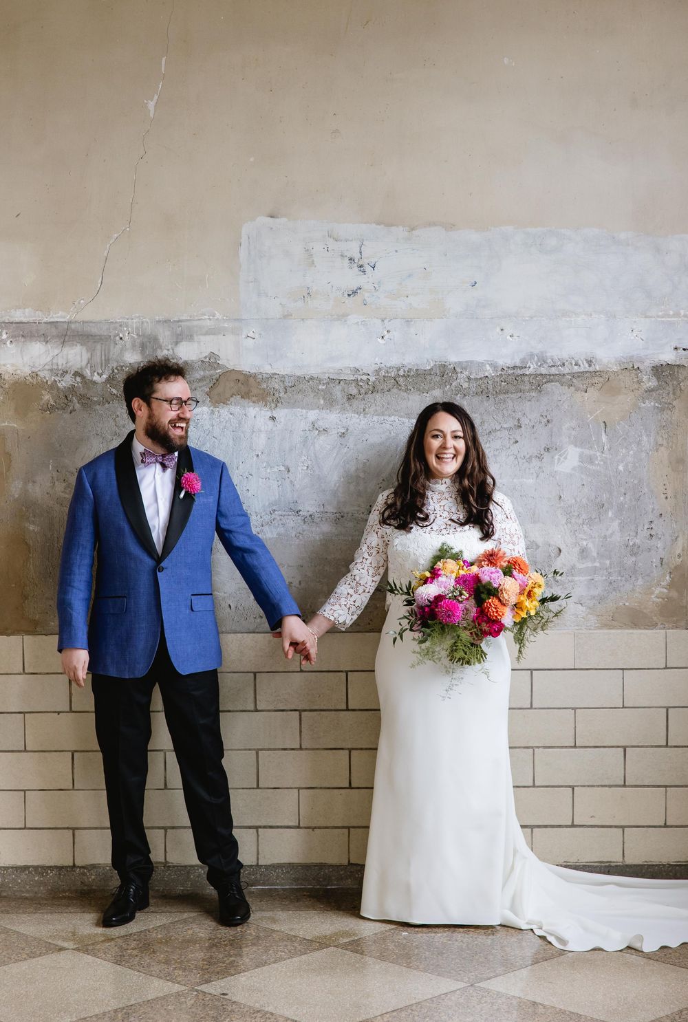 Wedding couple poses playfully against industrial wall with bride in white dress holding colorful bouquet. Bok Building Philadelphia