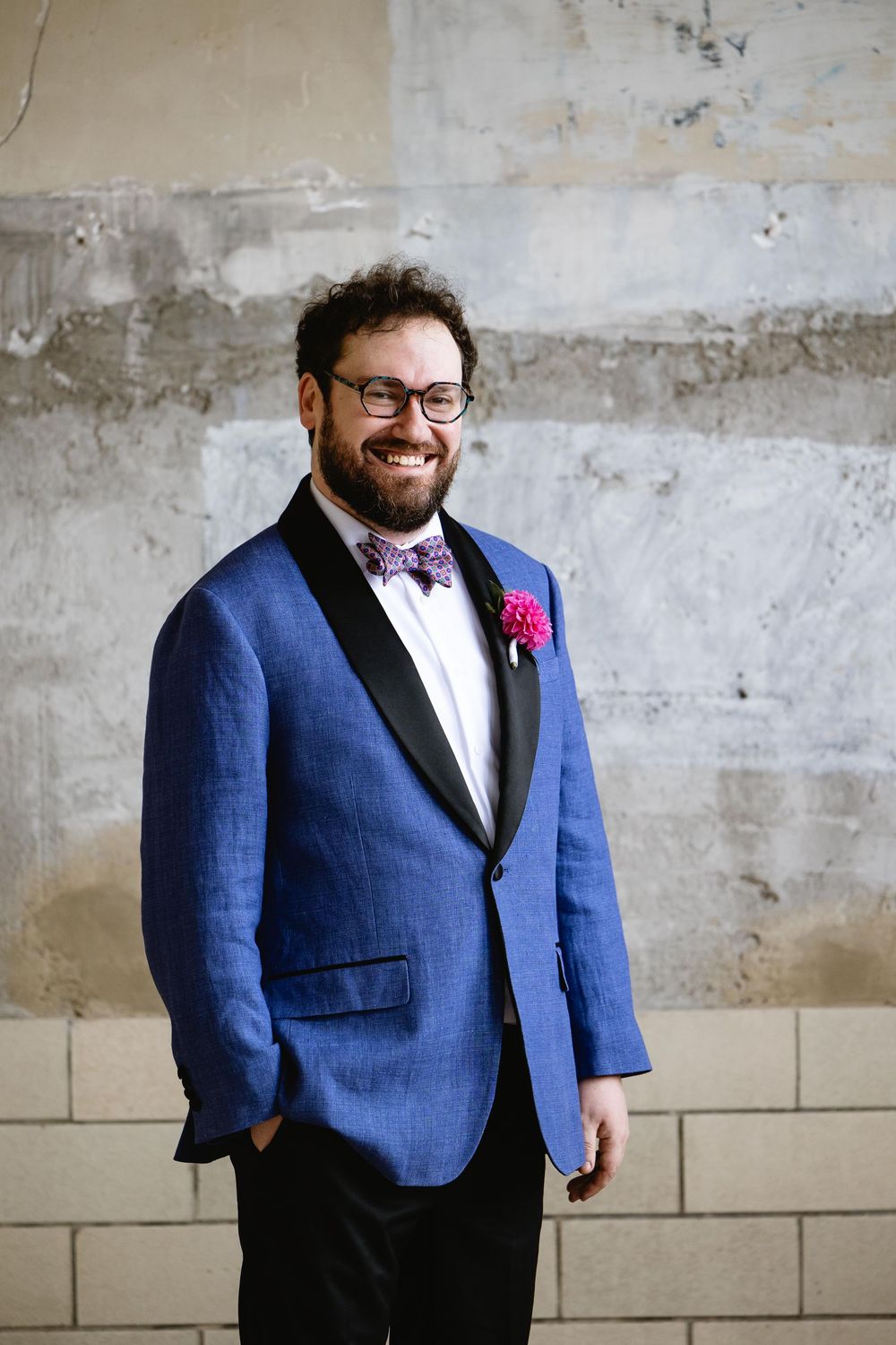 Groom in blue blazer and glasses poses against textured concrete wall with pink boutonniere. Bok Building Philadelphia