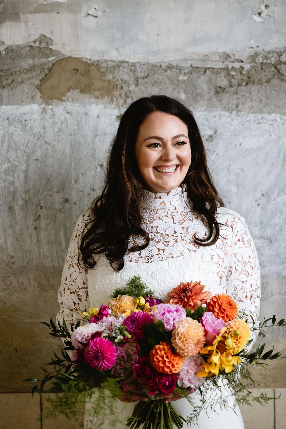 Bride in lace dress holds vibrant bouquet of pink, orange and yellow flowers against concrete backdrop. Bok Building Philadelphia
