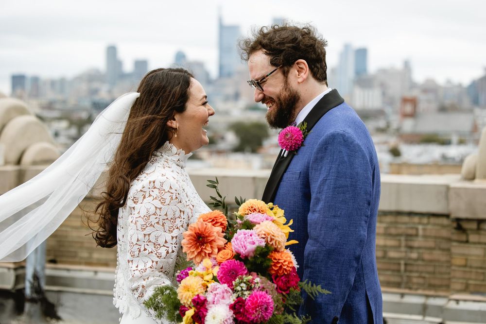 Couple poses for wedding photos on a rooftop with vibrant floral bouquet and city skyline backdrop. Bok Building Philadelphia