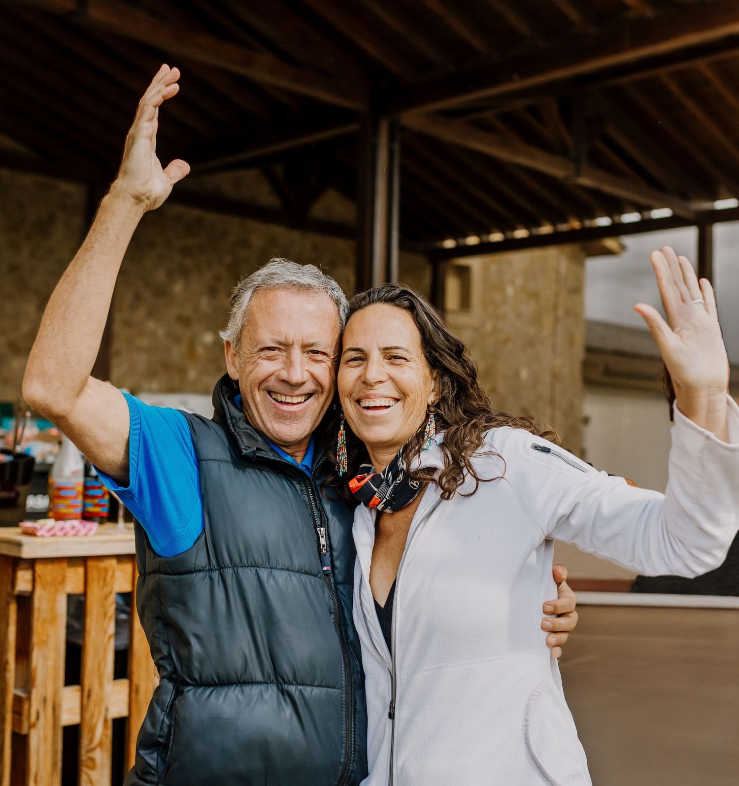 A happy couple hugging and lifting one hand each up, looking in the camera.