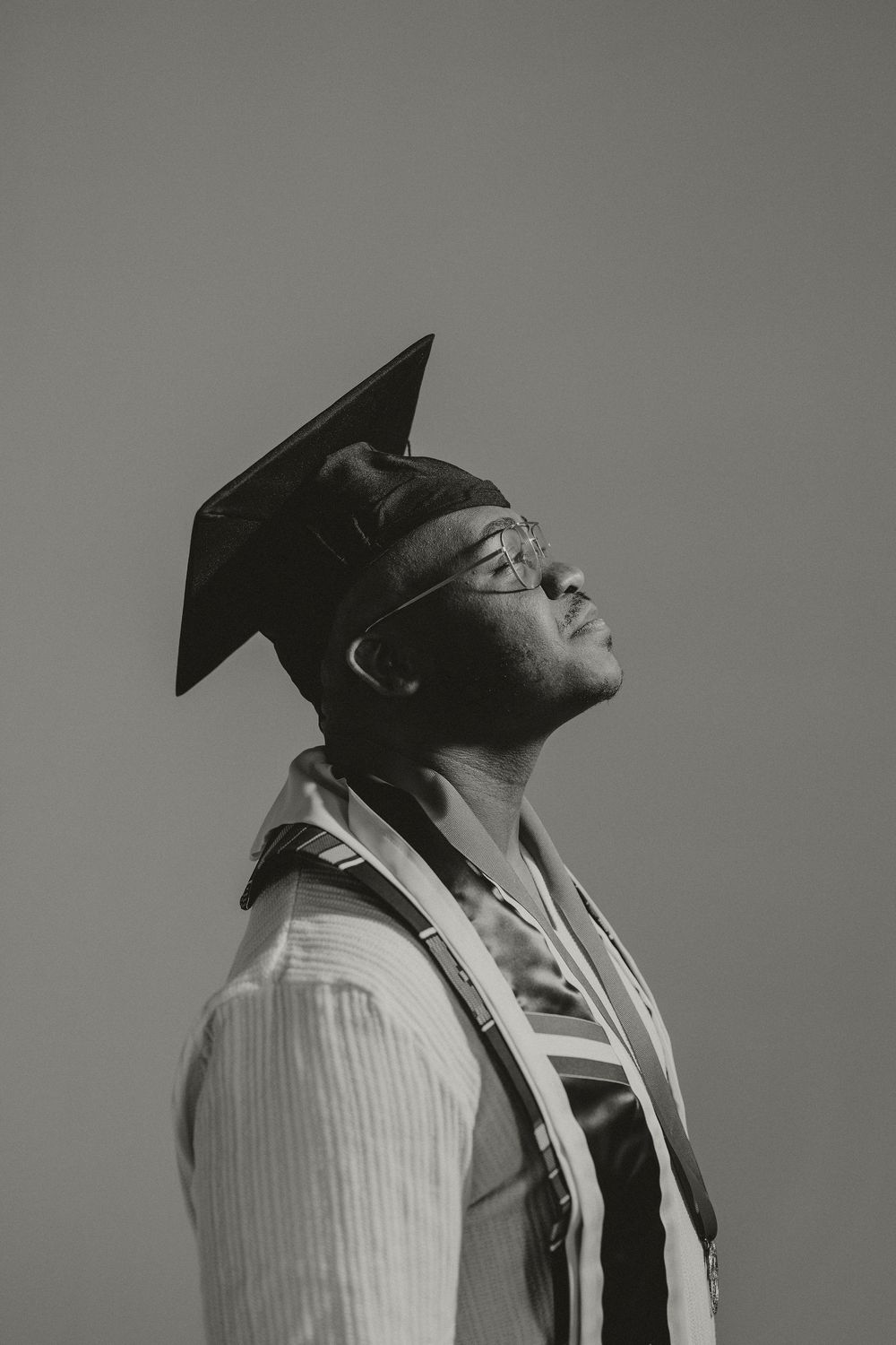 Black and white portrait of a graduate in cap and gown looking upward with contemplative expression.