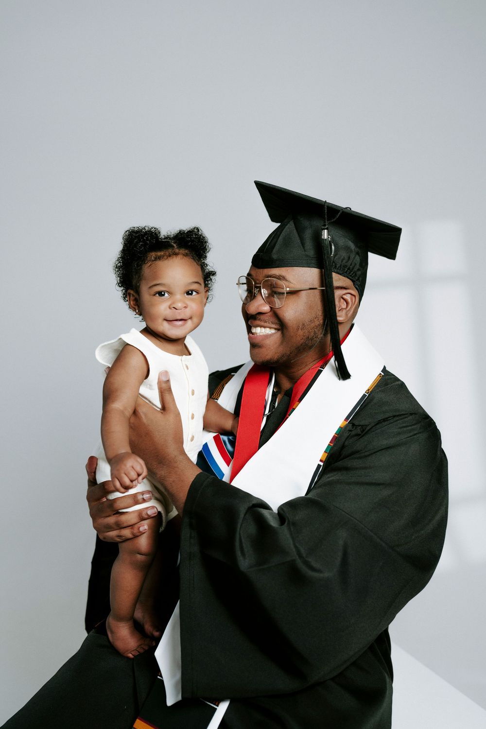 A graduate in cap and gown shares joyful moments with a small child against a white backdrop.