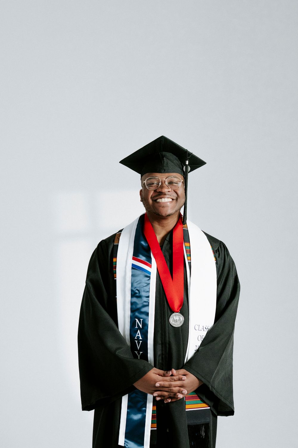 Graduate in academic regalia smiles while wearing graduation cap, gown, and colorful stoles against white background.