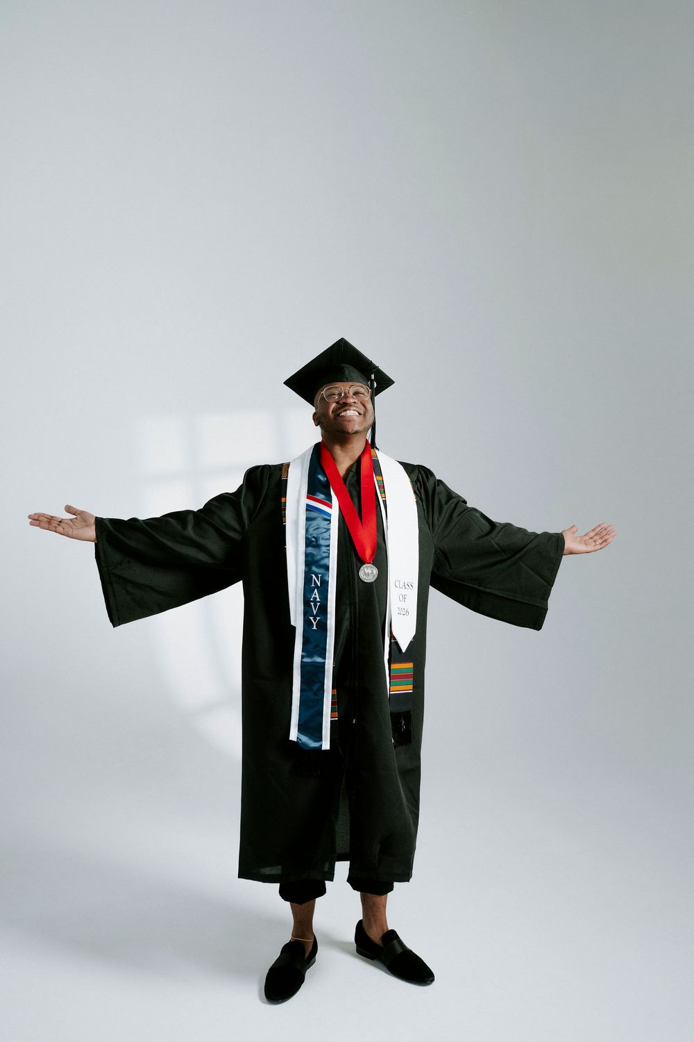 Graduate in academic regalia spreads arms wide in celebration against light background.
