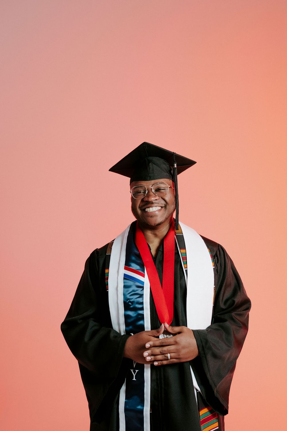 Graduate in black cap and gown poses confidently against a pink background.