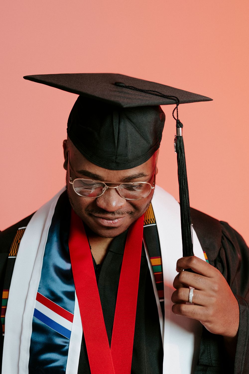 Close-up graduation portrait showing graduate in cap, gown and stoles against coral background.