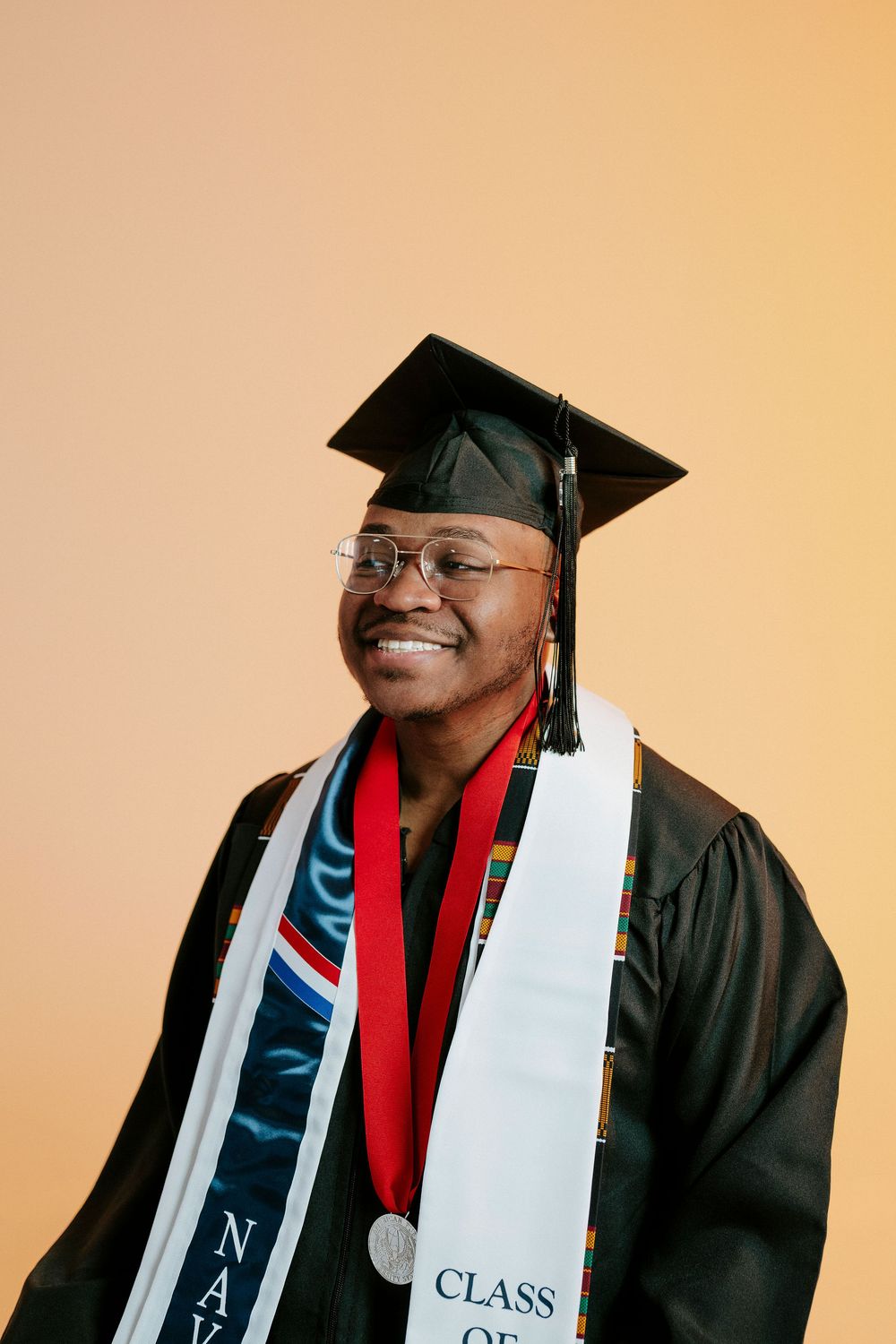 Graduate in cap and gown wearing graduation stole and honor cords poses for portrait against warm background.