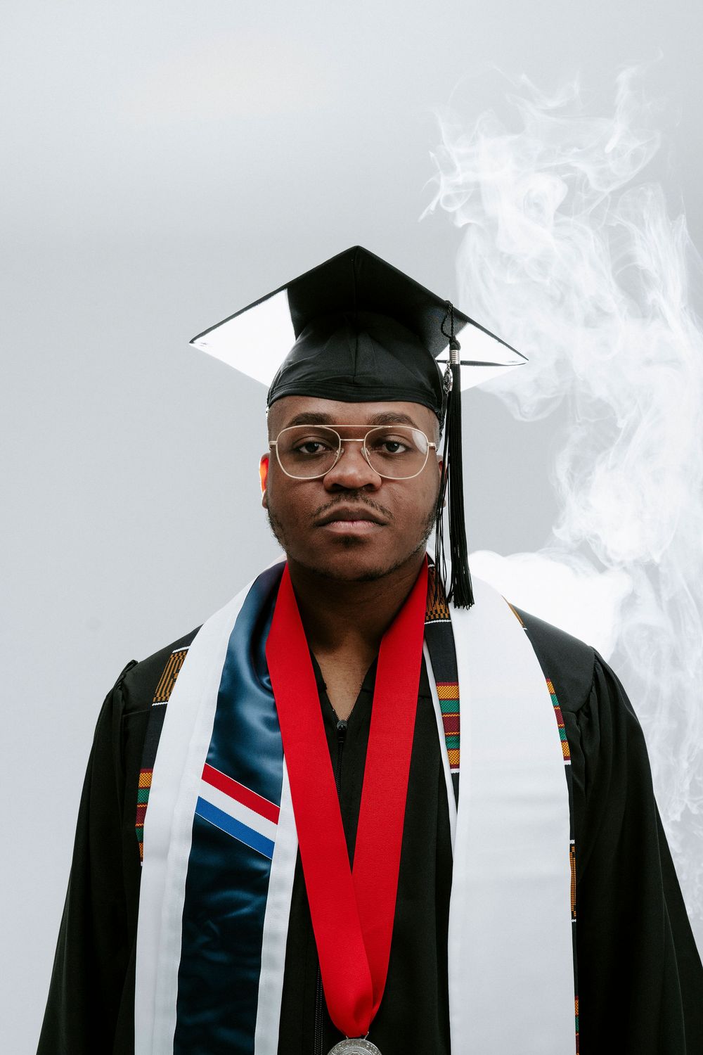 Graduate in academic regalia poses with smoke effect against grey background.