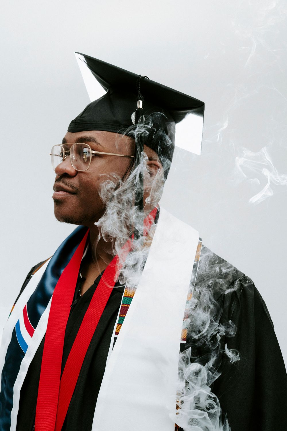 Graduate wearing a black cap and gown with red stole exhales smoke in an artistic photo series.