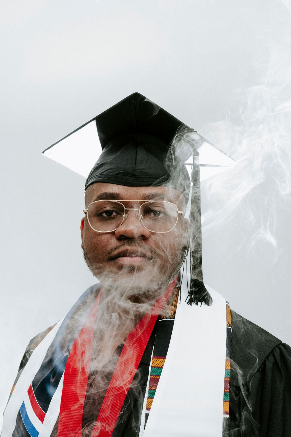 Graduate in academic regalia and glasses poses with smoke effects against light background.