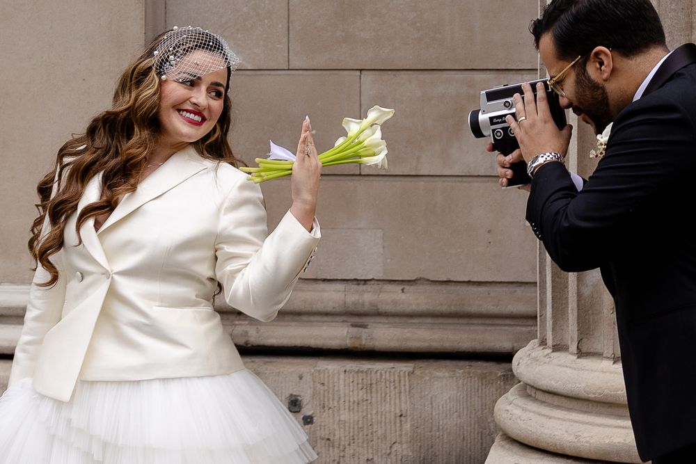 A photographer captures a person in a white wedding outfit posing with a bouquet outside a classical stone building.