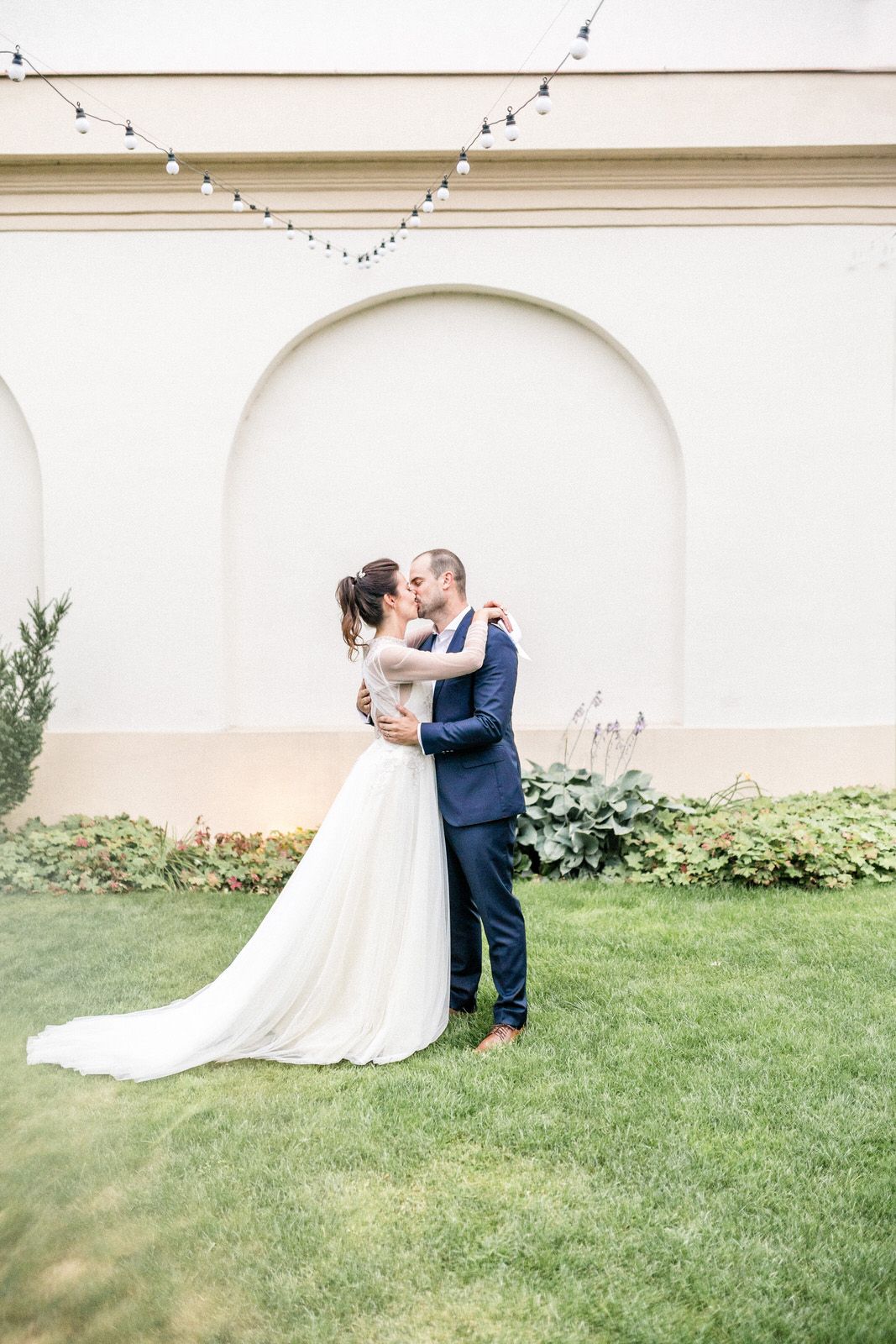 Newlyweds sharing romantic moment under white archway with string lights.