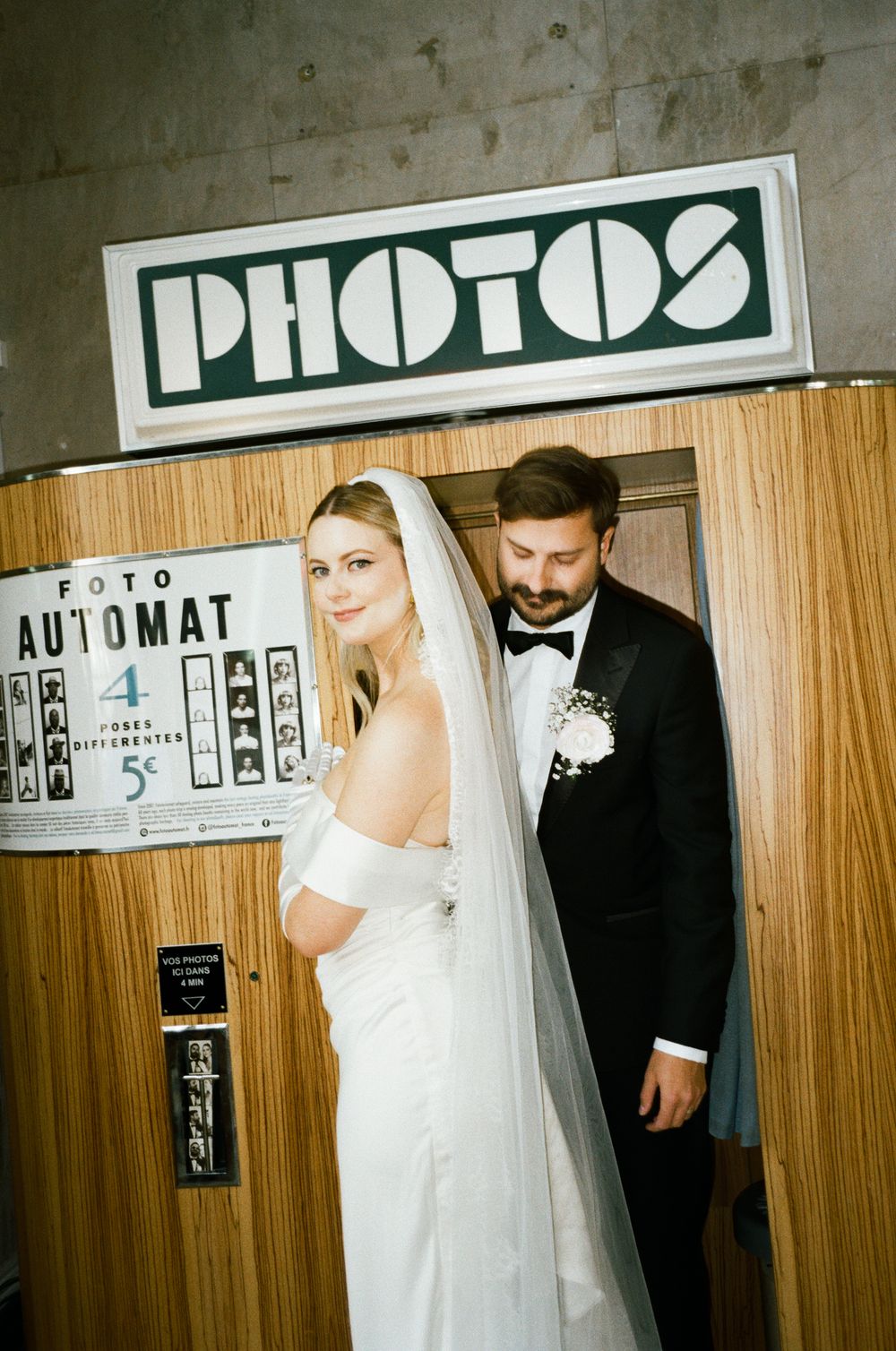 A couple in wedding attire poses in front of a vintage photo booth with wood paneling and signage.