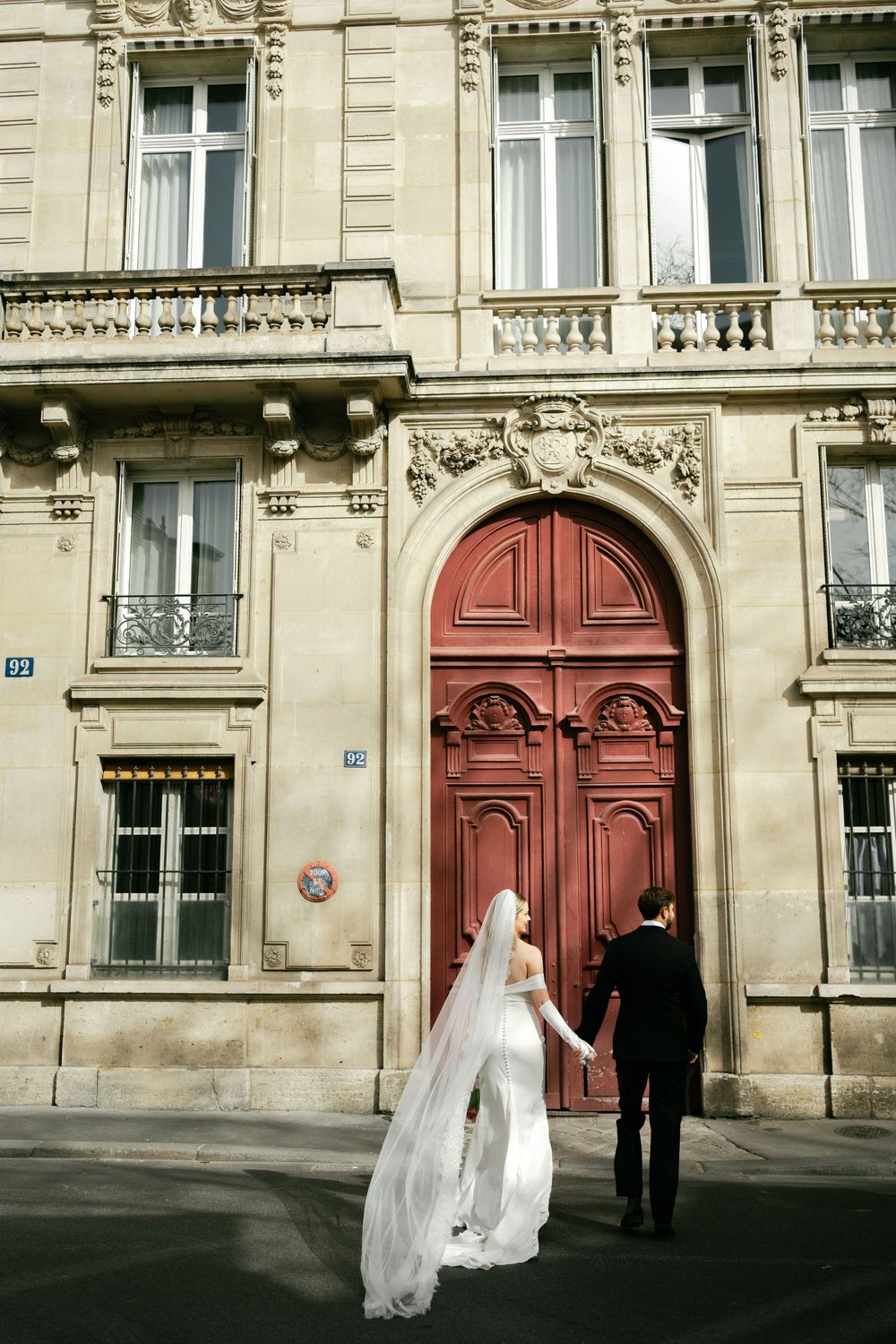 A couple in formal attire stands before a grand red door on an ornate Parisian building facade.