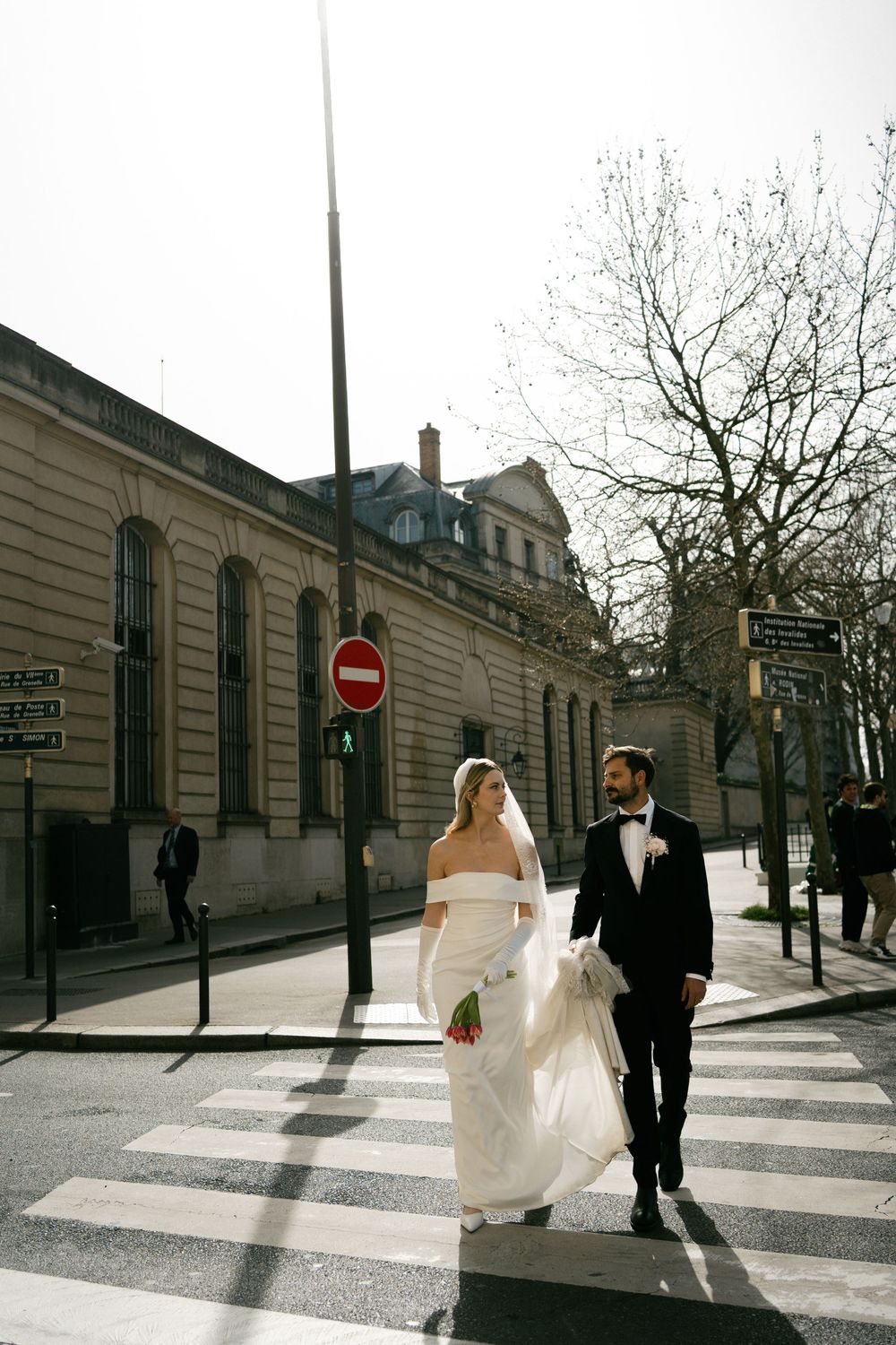 A couple in wedding attire walks across a crosswalk in front of historic buildings on a bright sunny day.