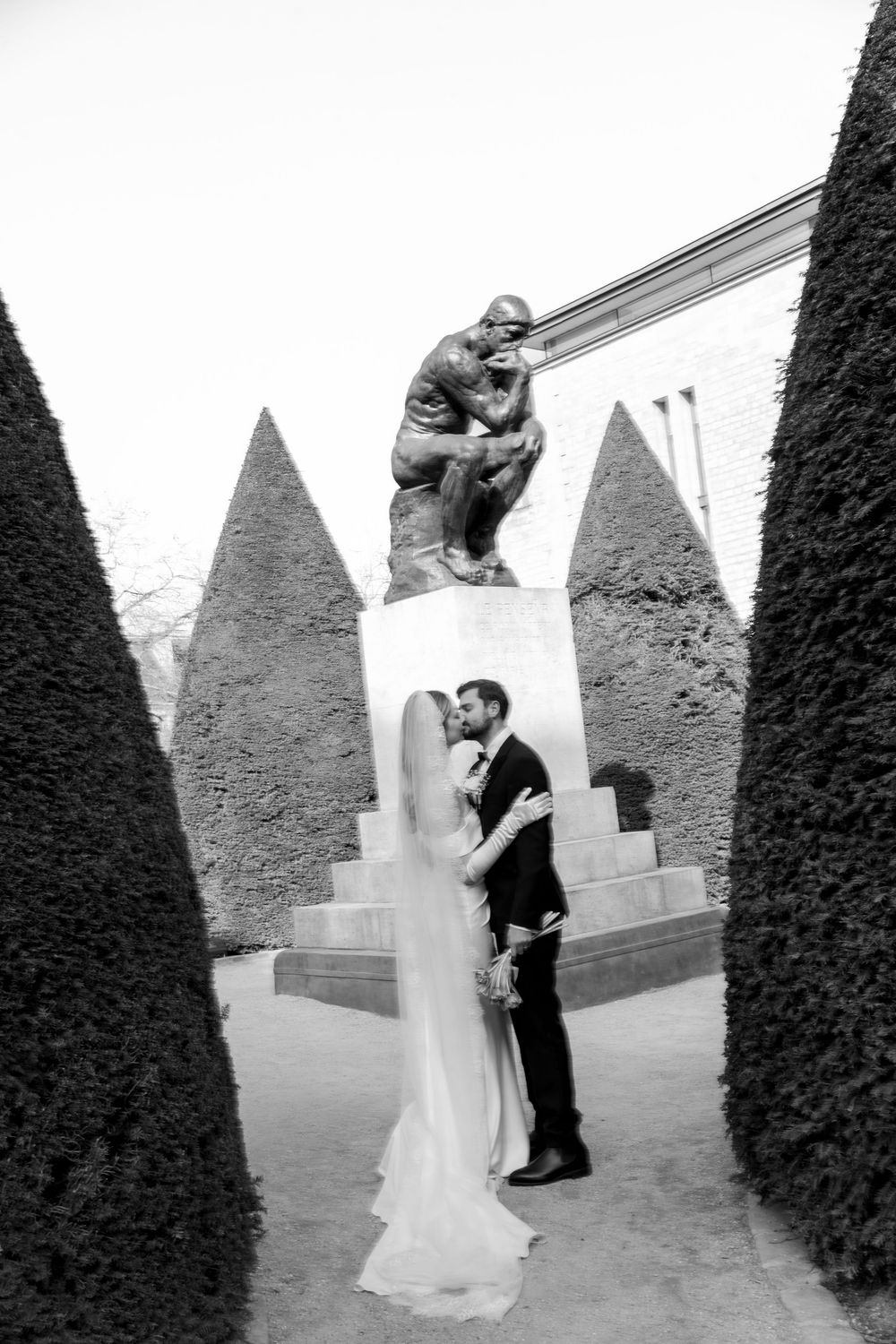 A black and white wedding photo series showing a couple embracing among geometric topiary in a formal garden setting.