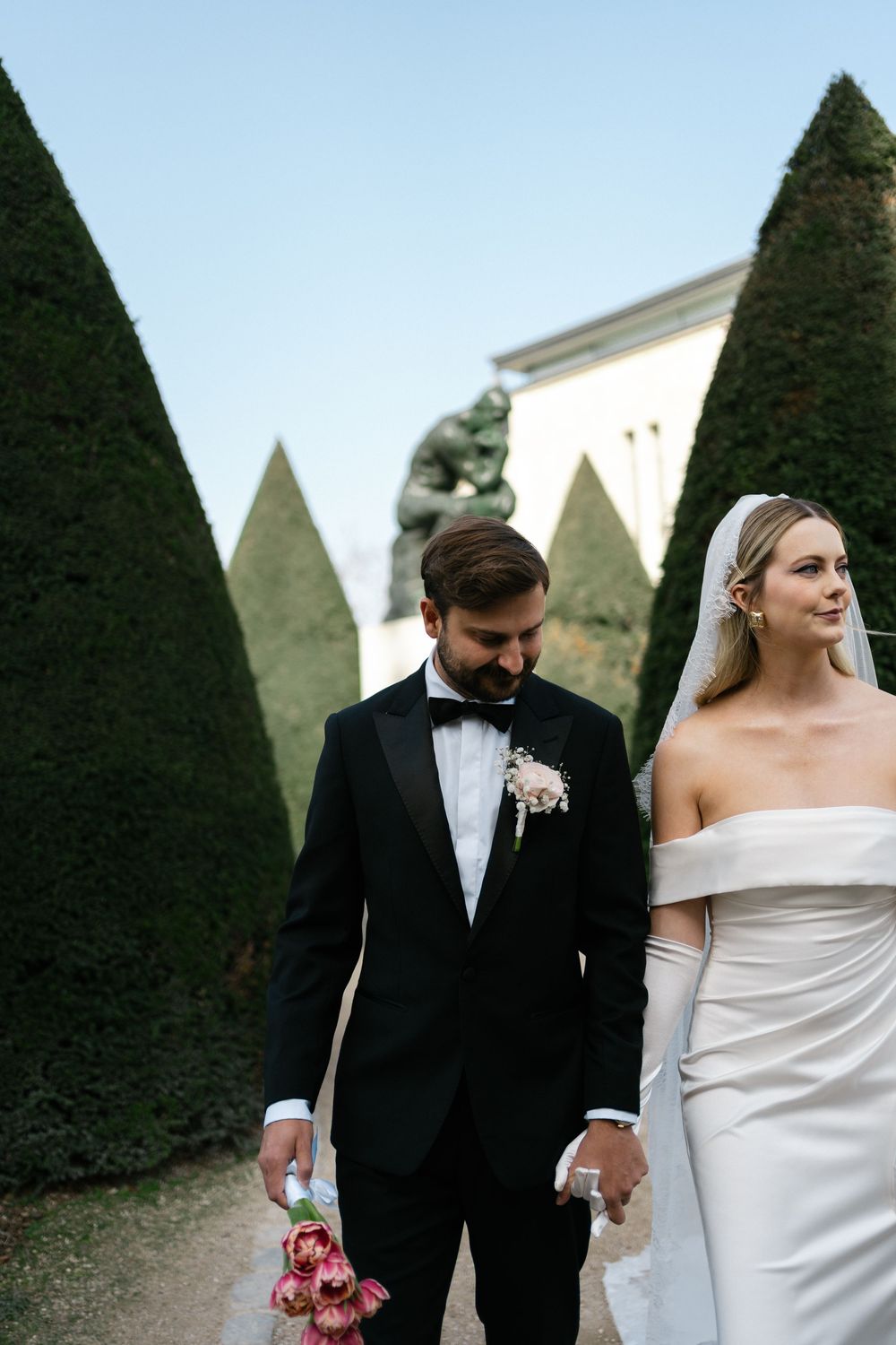 A couple in formal wedding attire walking through a garden with manicured topiary hedges in the background.