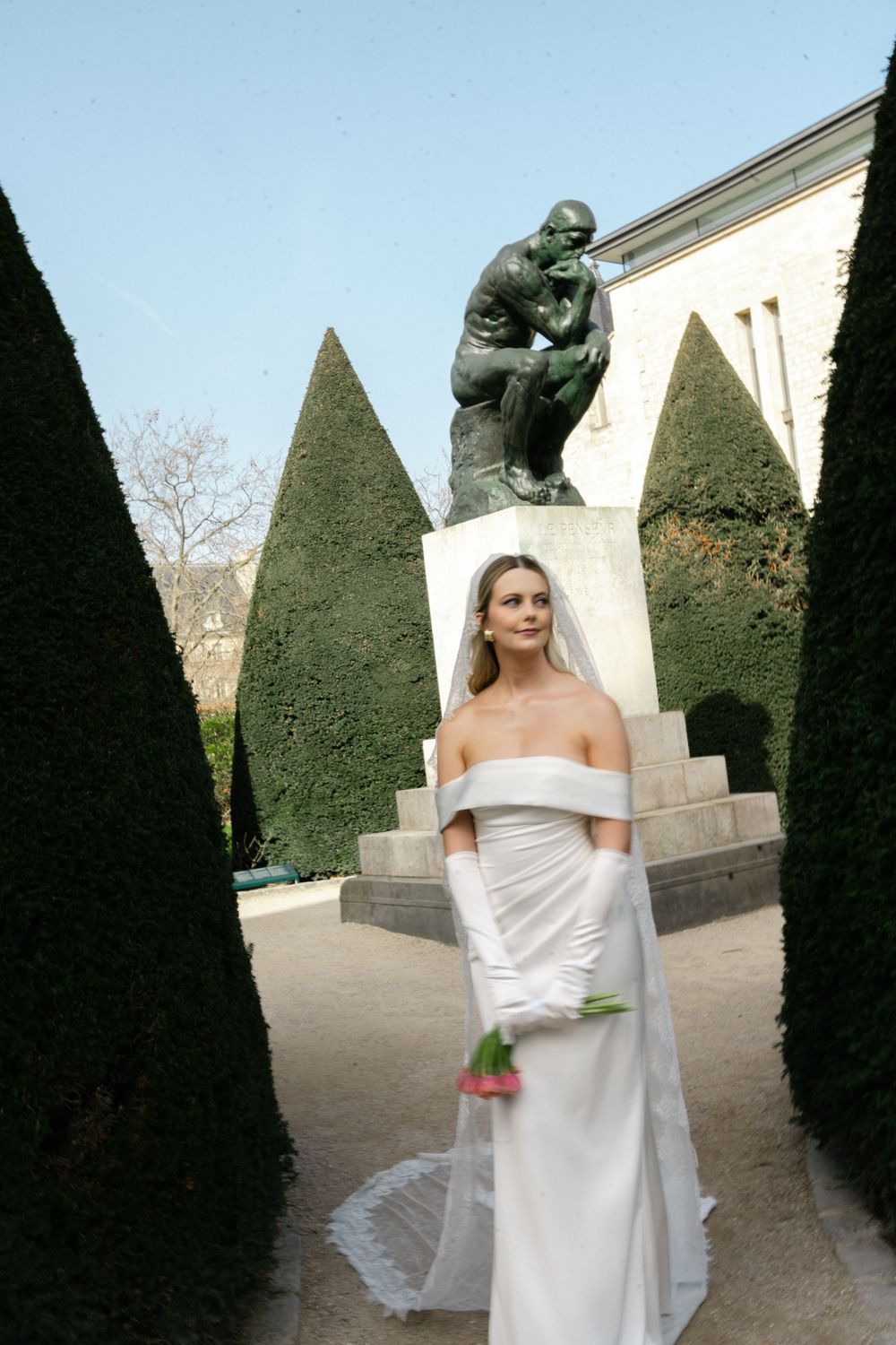 Woman in white dress poses artistically in formal garden with sculpted hedges and classical statues.