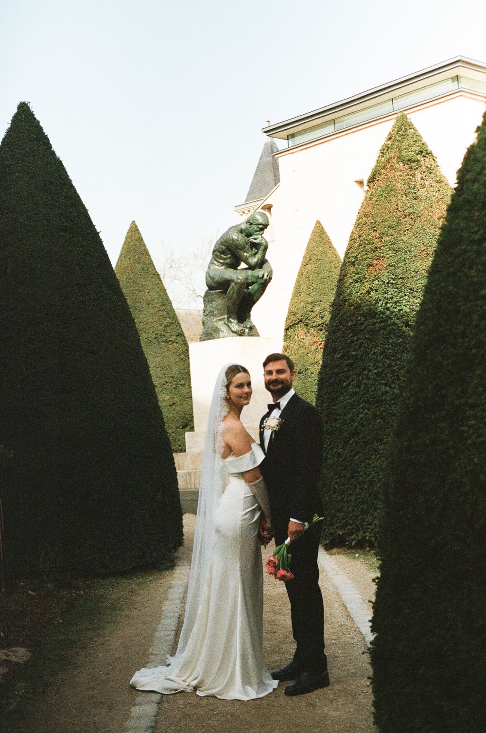 A couple poses between tall manicured hedges in a formal garden setting during their wedding photoshoot.