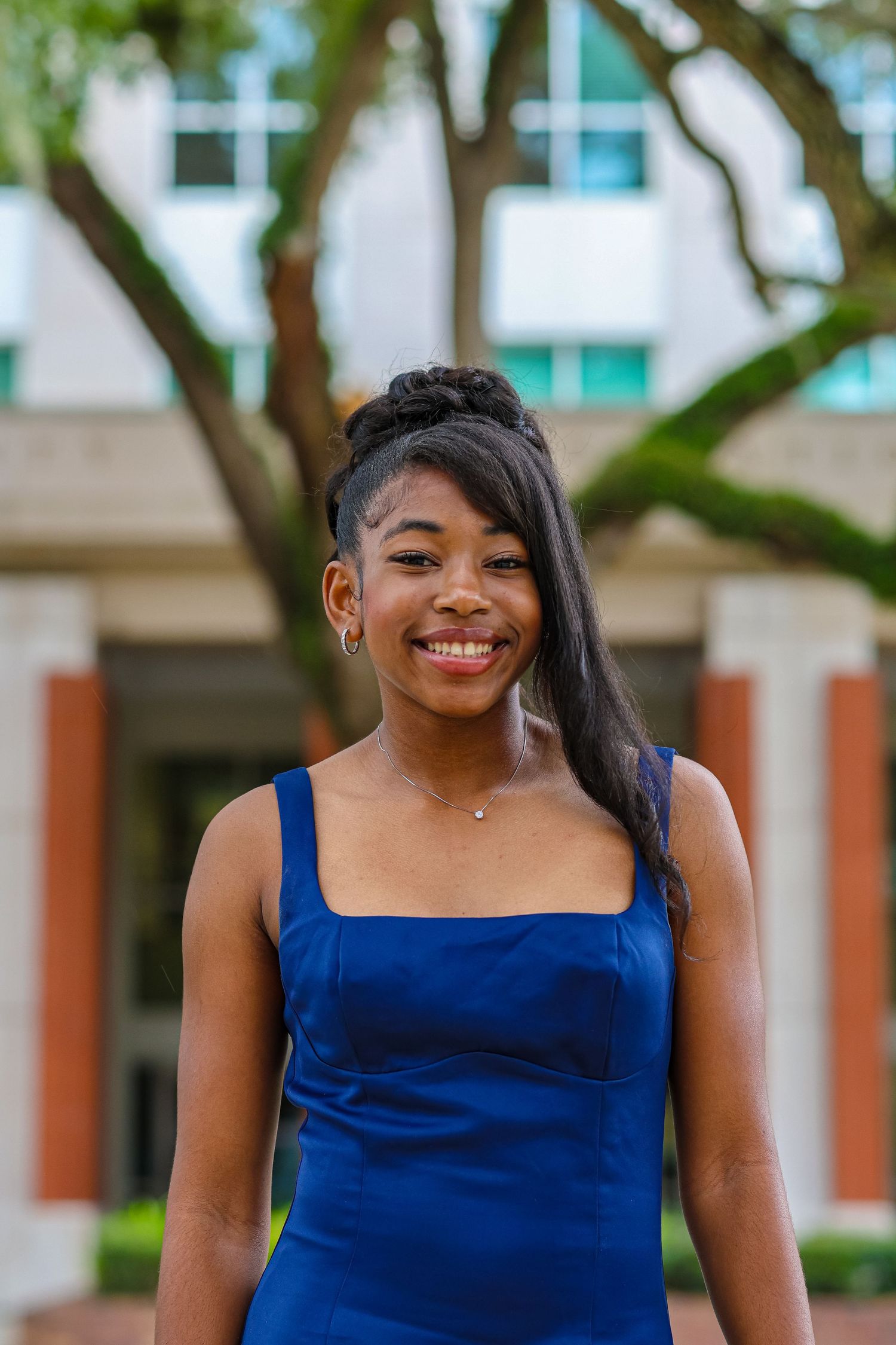 Person in a royal blue sleeveless dress smiles warmly while standing outdoors near a building.