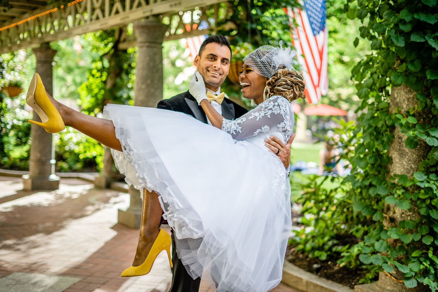 A joyful wedding couple shares a romantic moment outdoors with an American flag in the background.