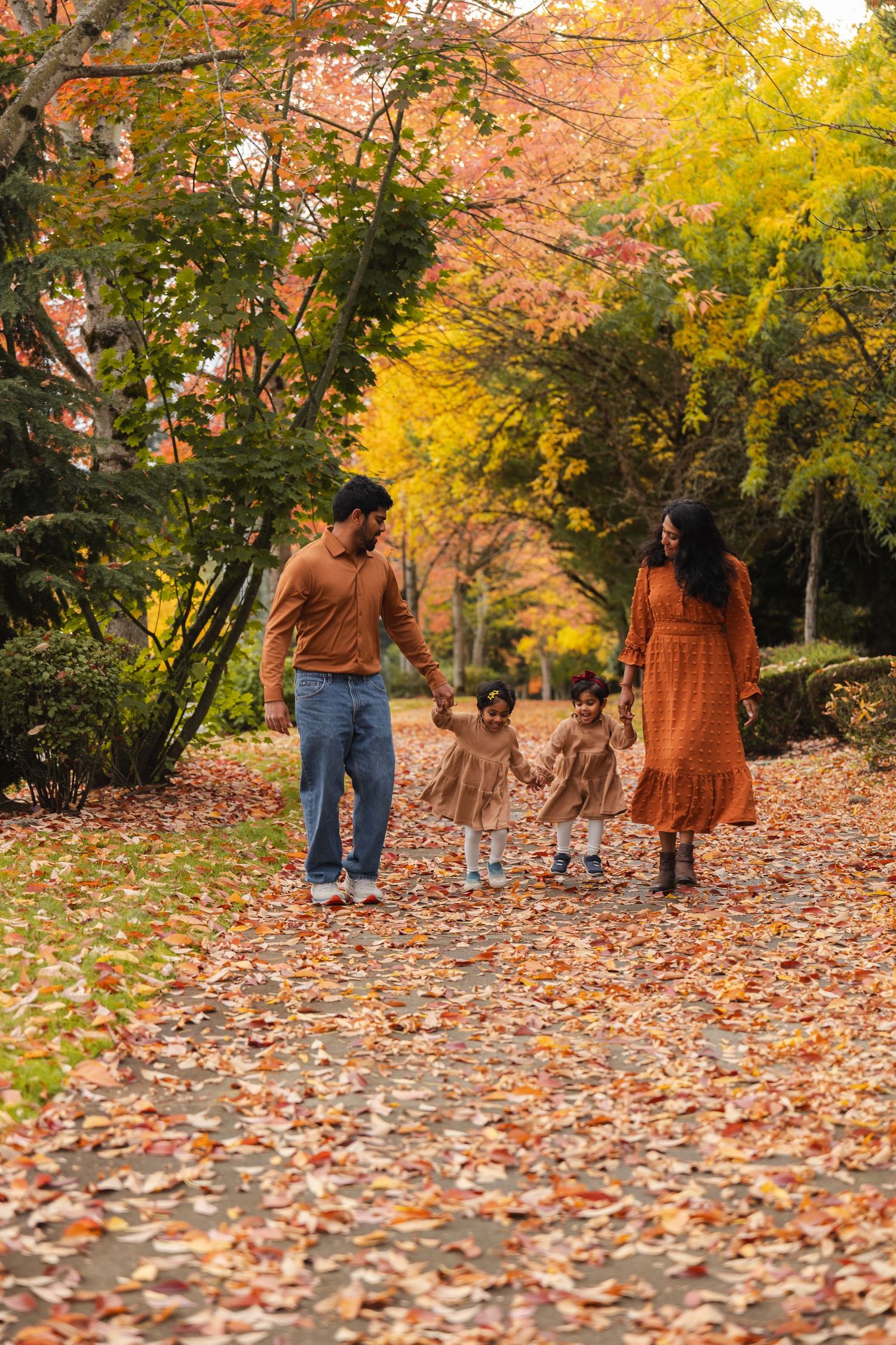 A family walks together on a leaf-covered path surrounded by autumn trees with vibrant orange and yellow foliage.