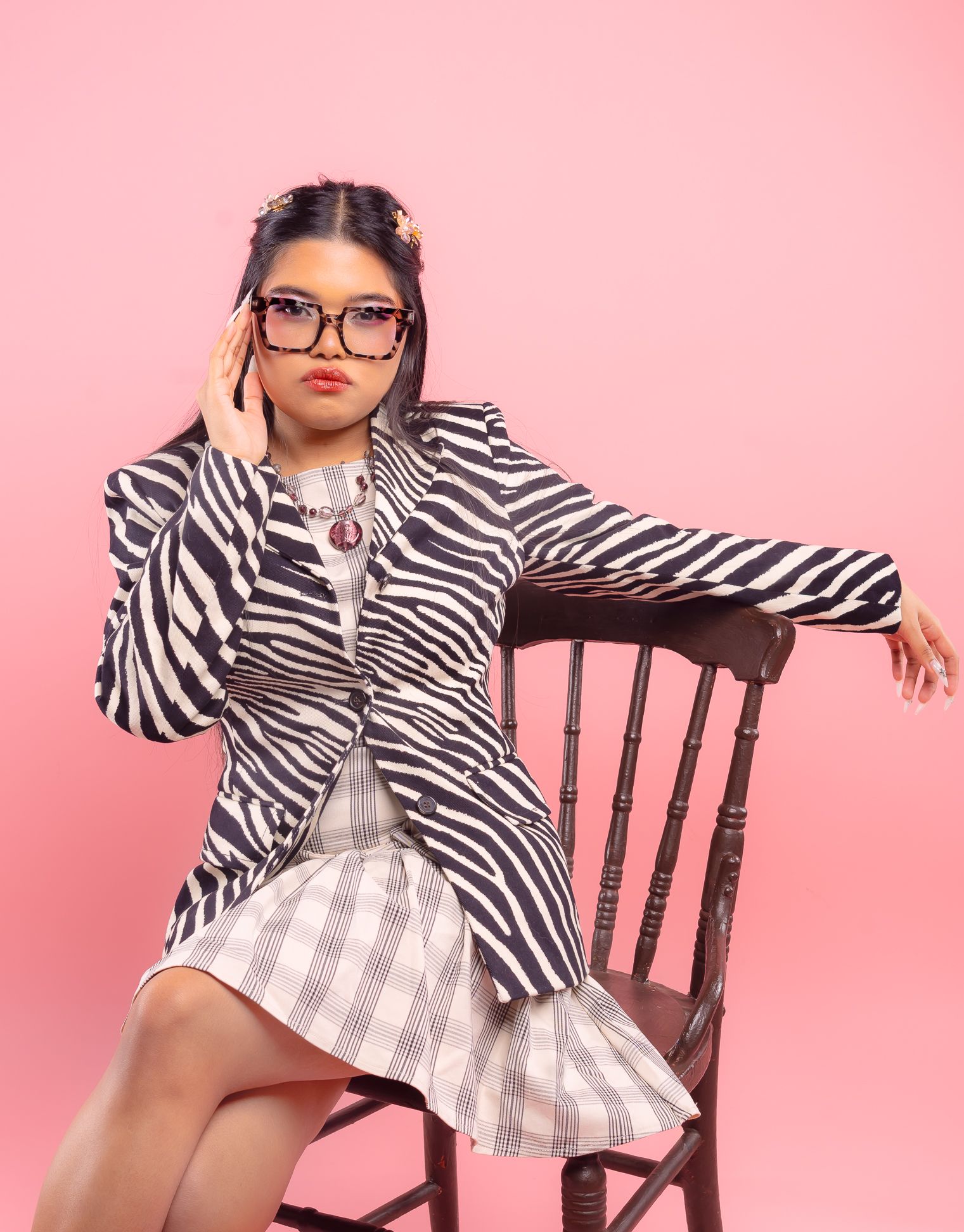 Girl in front of a pink backdrop, wearing a zebra-patterned blazer, beige-plaid dress and glasses, sitting on a chair looking straight