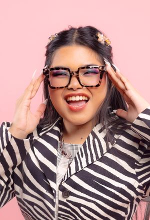 Girl in front of a pink backdrop, wearing a zebra-patterned blazer, mouth open with a happy smile, touching glasses with hands