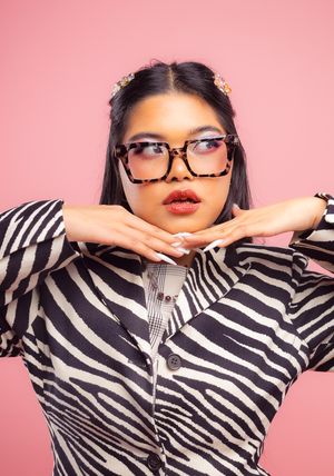 Girl in front of a pink backdrop, wearing a zebra-patterned blazer, hands under chin, looking to the right