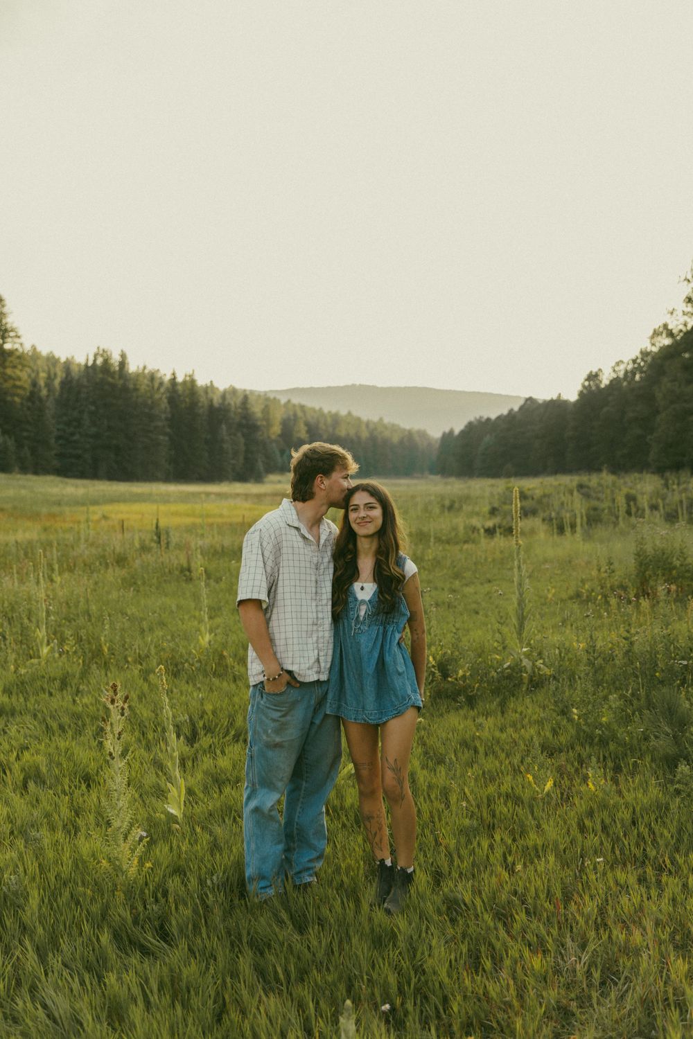A romantic couple stands together in a scenic mountain meadow at golden hour.
