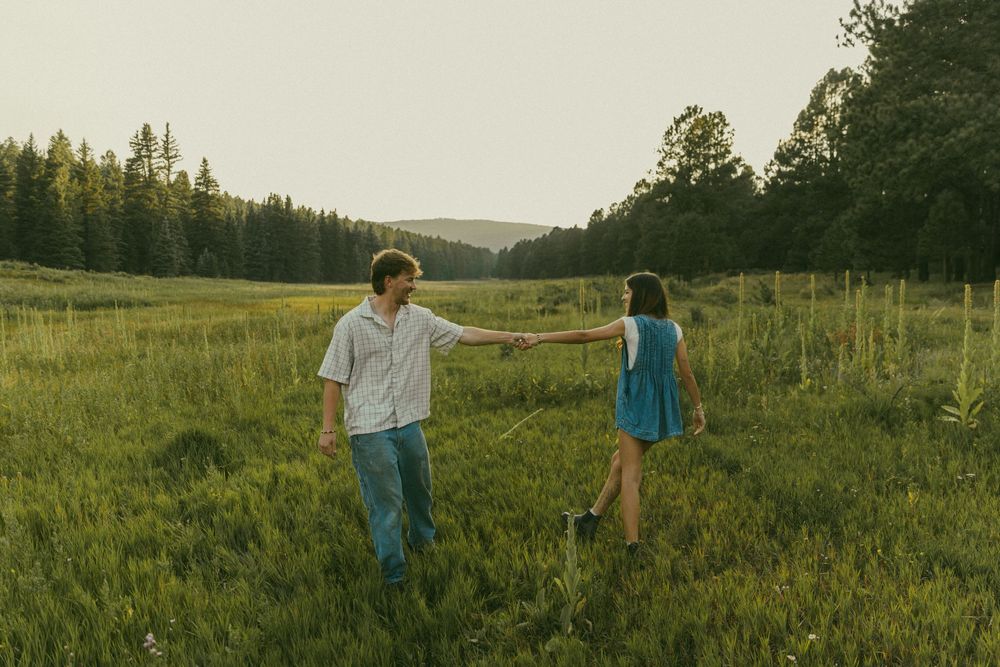 A couple stands together in a golden meadow at sunset with mountains and forest in the background.