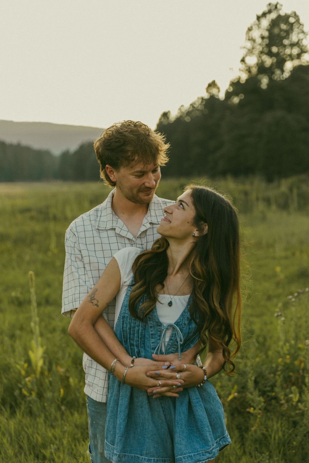 A romantic couple embraces in a sunlit meadow with rolling hills and trees in the distance.