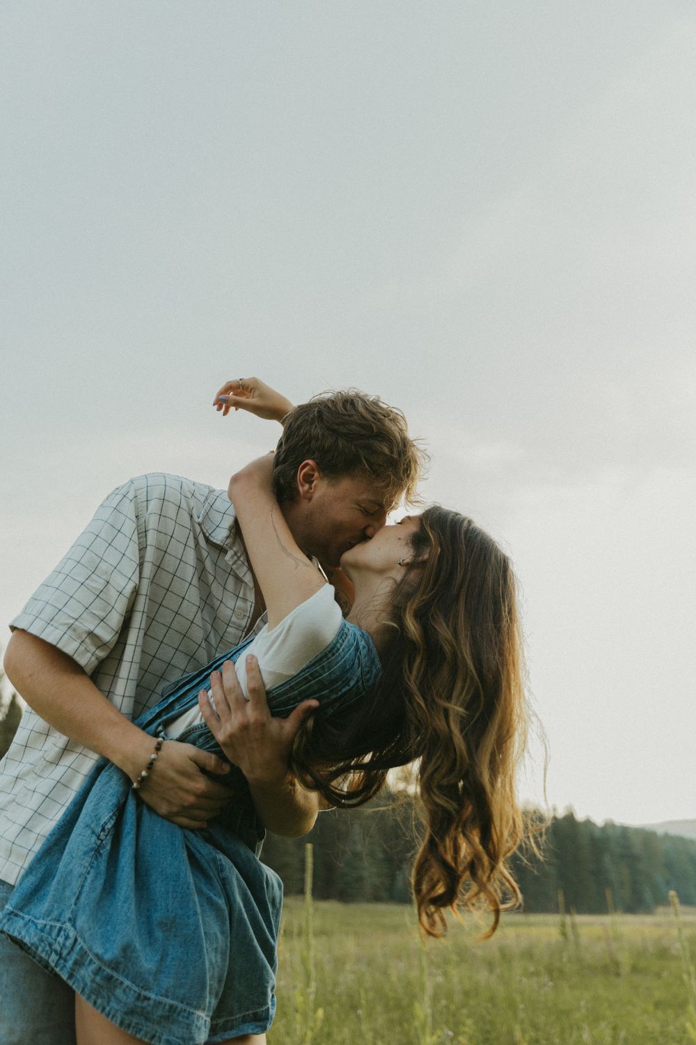 A romantic couple shares a passionate kiss while standing in a lush green meadow on a cloudy day.