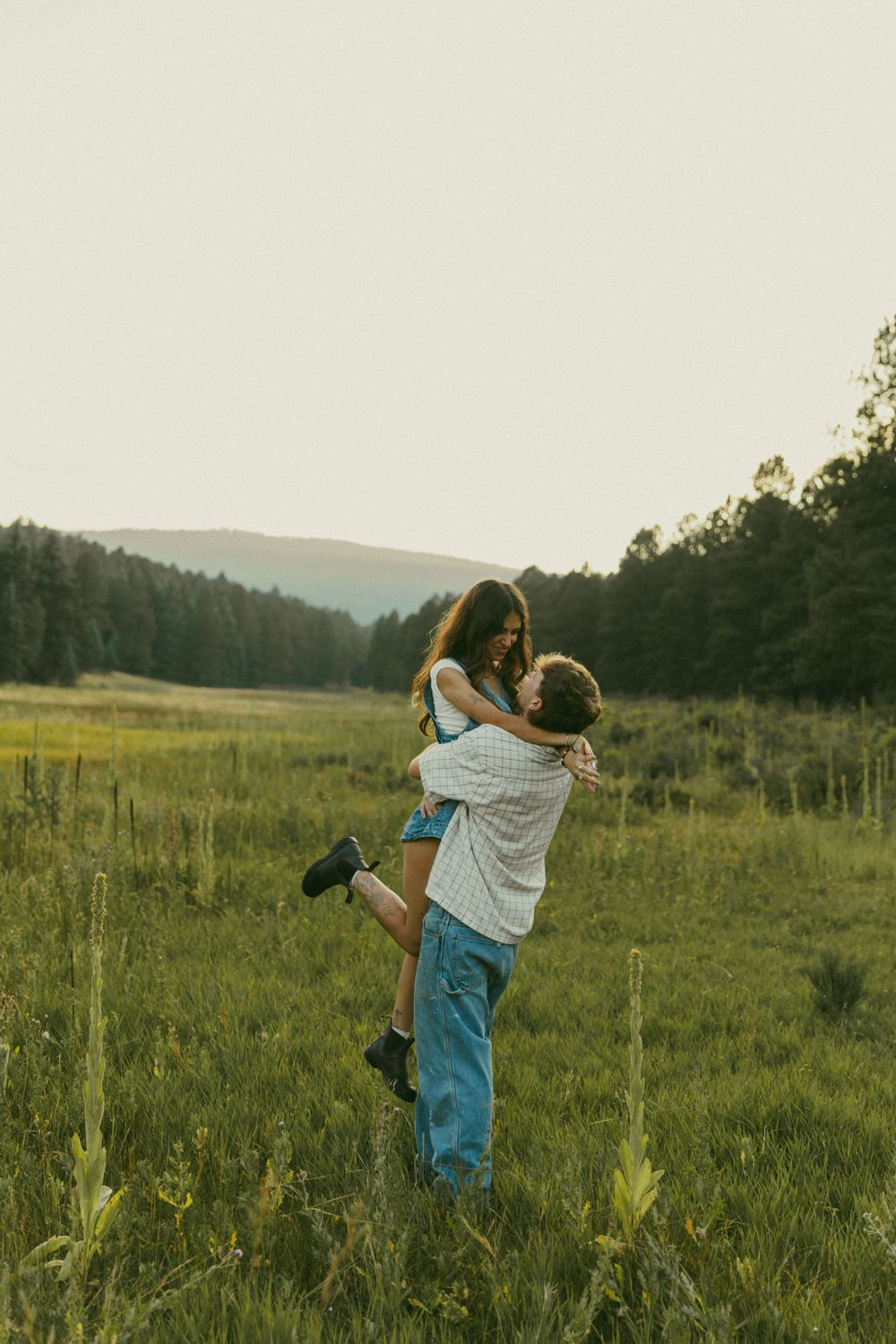 A joyful couple shares a tender moment in a grassy field with forest and mountains visible in the distance.