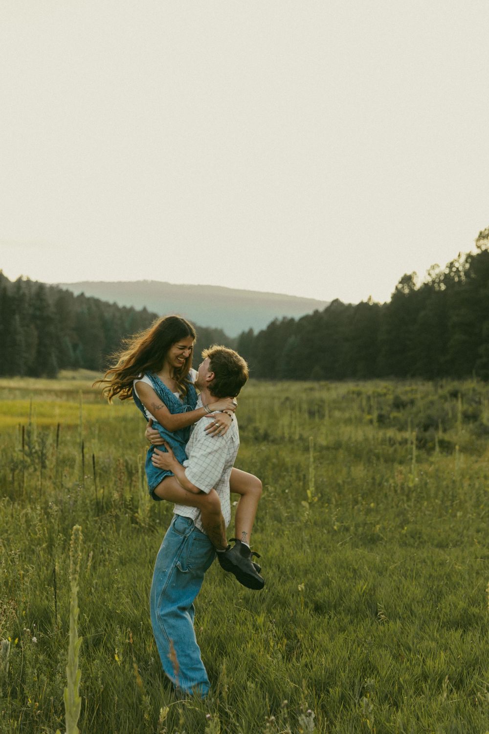 A playful couple shares a joyful moment in a mountain field during sunset.