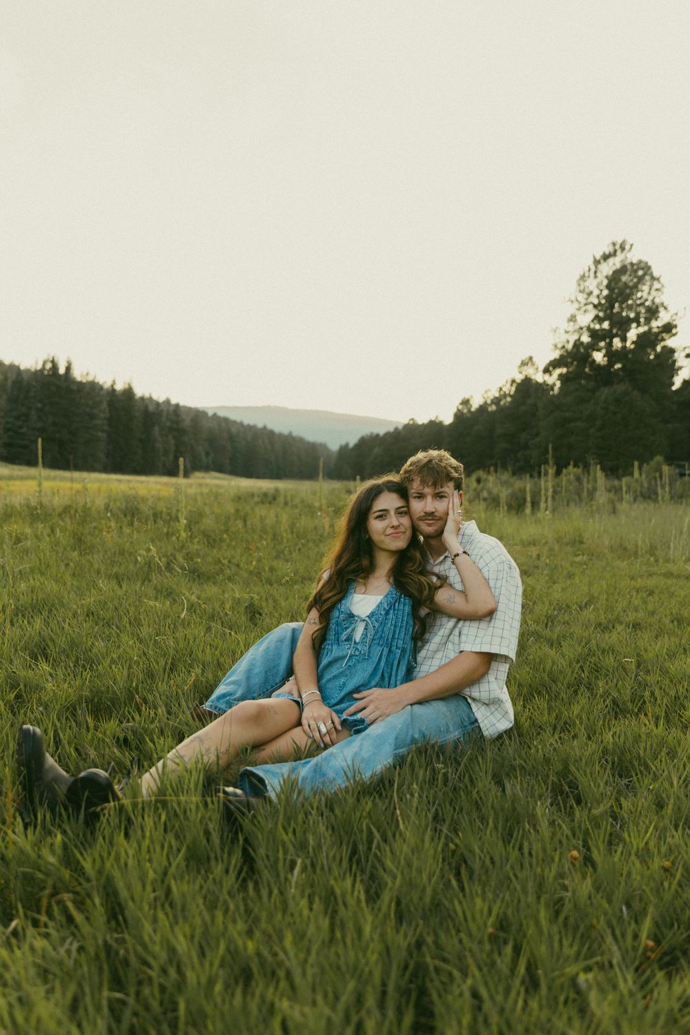 A couple sits together in a serene meadow at sunset with mountains and forest in the background.