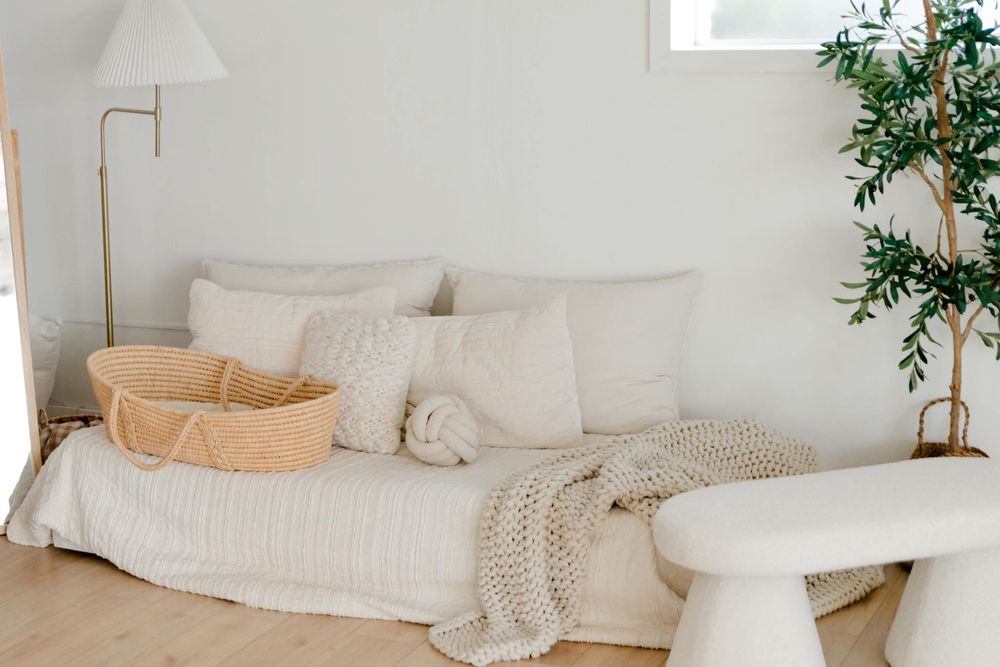 Minimalist nursery with a cream daybed, chunky knit blanket, wicker Moses basket, floor lamp, and potted olive tree.