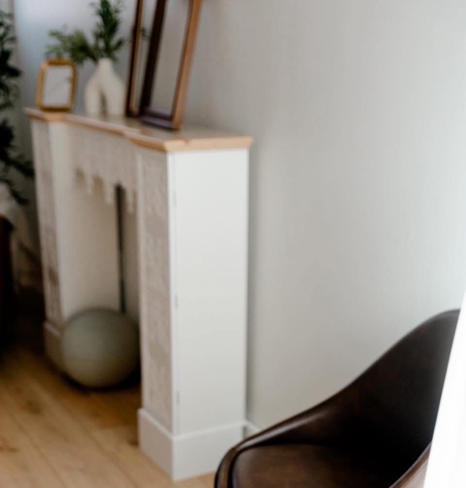 Modern white decorative fireplace mantel with wooden top, framed art, and a brown leather chair on hardwood floors.