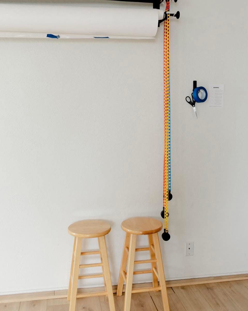 Two wooden bar stools beneath a wall-mounted paper roll backdrop with colorful rope pull in a studio.