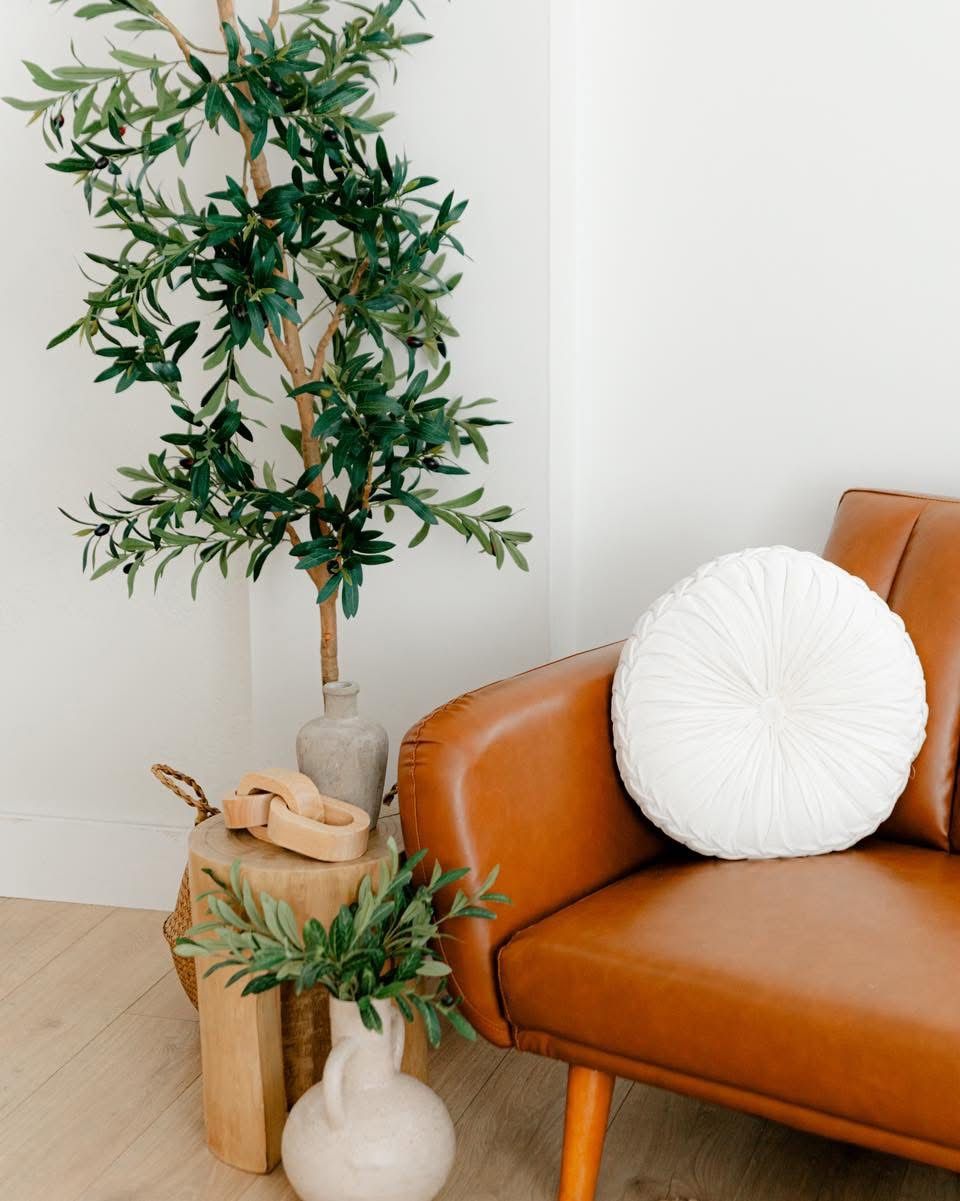 Tan leather armchair with white round pillow beside tall olive tree and small white vase in bright minimalist living room.